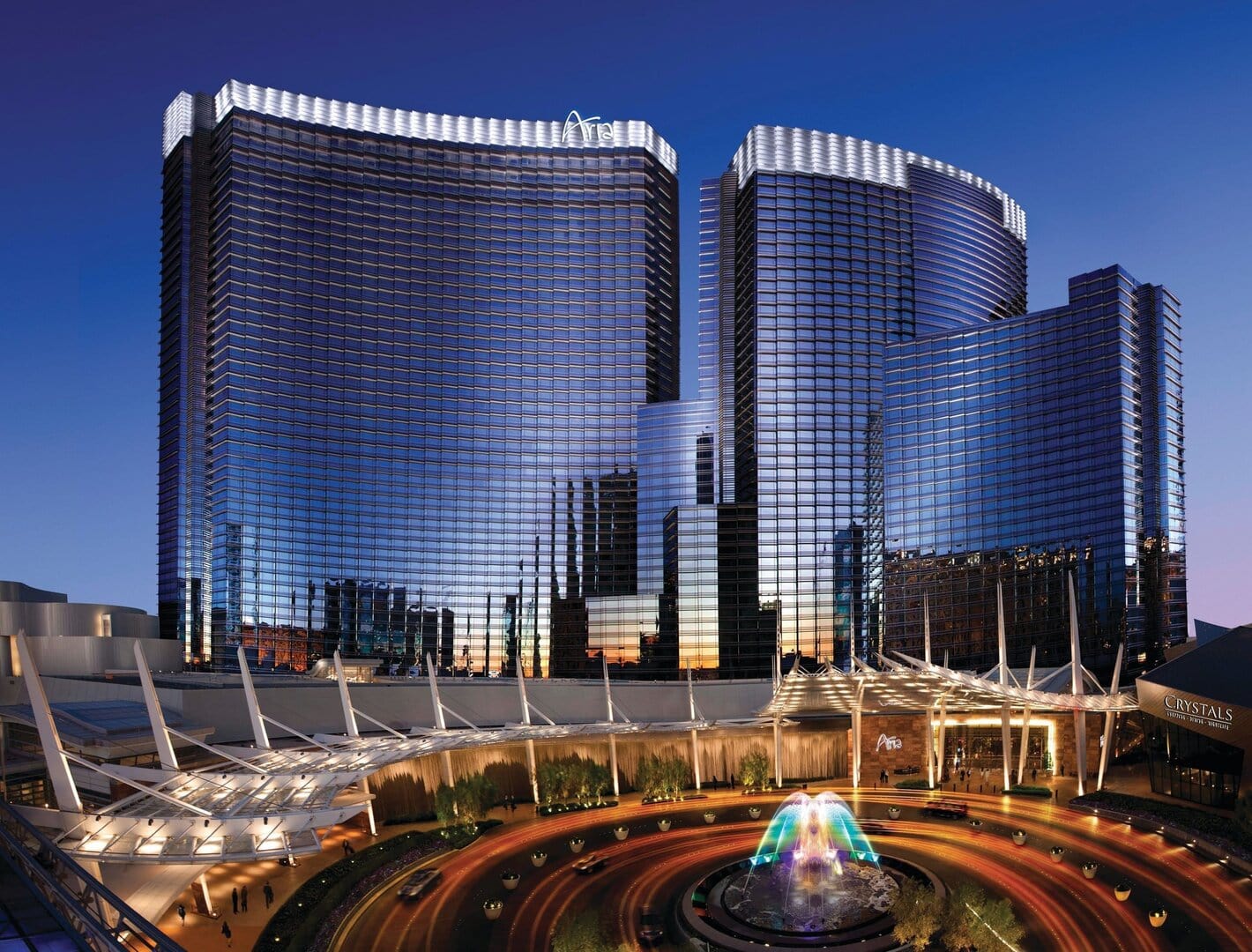 Large dark blue, glass built building, with white at the highest point with text saying: Aria, and a fountain with colourful waters.