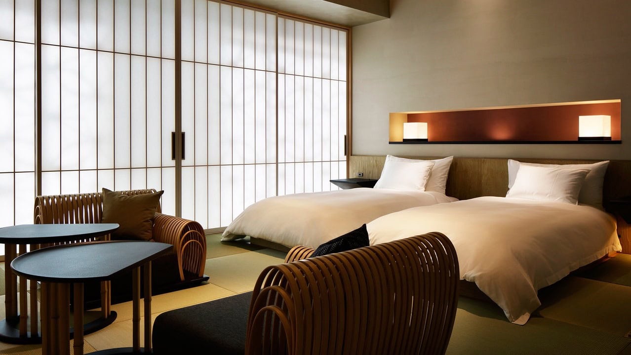 Two single beds with white decor, night lamps over bed, and seating area with beige chairs and black tables.